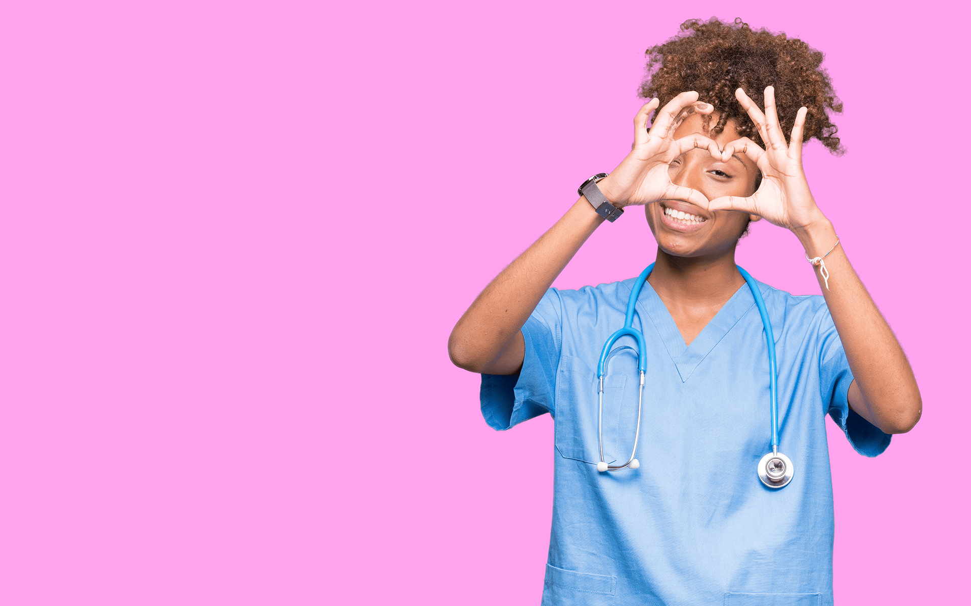 3 Ways Your Emotions Can Support Your Heart Health—Image of a Black female healthcare worker wearing blue scrubs and a stethoscope holds her hands up in front of her face and makes a heart shape with her fingers. She stands in front of a pink background.