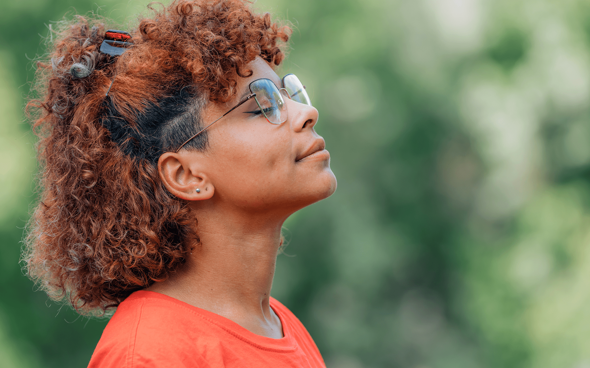 一位黑发卷曲、戴大眼镜、穿橙色衬衫的女人站在户外，脸朝上。她脸上带着平静的表情。