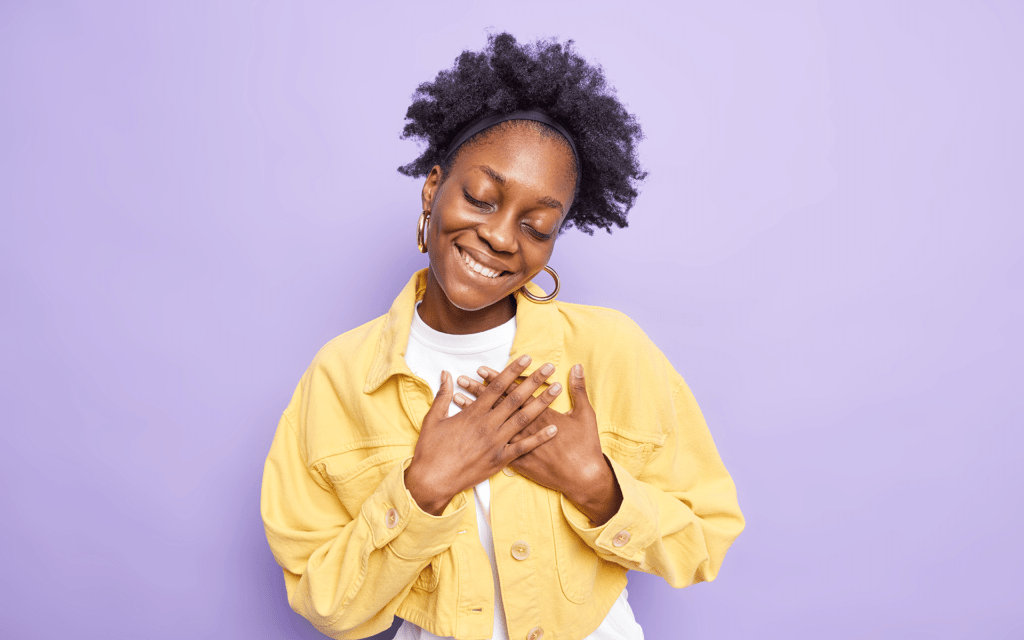 一位年轻的黑人女性微笑着，双手放在胸前，站在紫色工作室背景前。