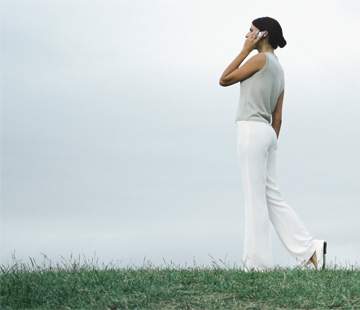 一名穿着无袖上衣和宽腿裤的女子站在户外草地上，对着手机说话。她侧身对着背景中的多云天空，短暂地停下来考虑是否需要断开连接。