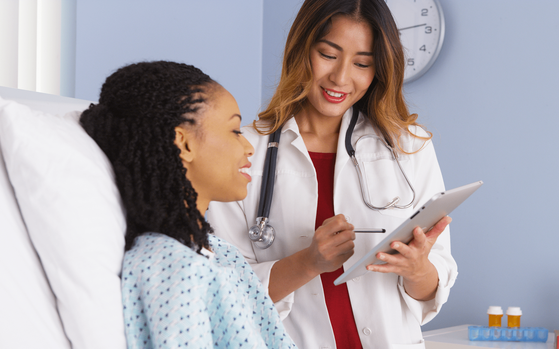 命名如何赋予患者权力及其他正念新闻——一位日本女性医生向坐在病床上微笑的黑人女性展示她的平板电脑内容。两人都在微笑。