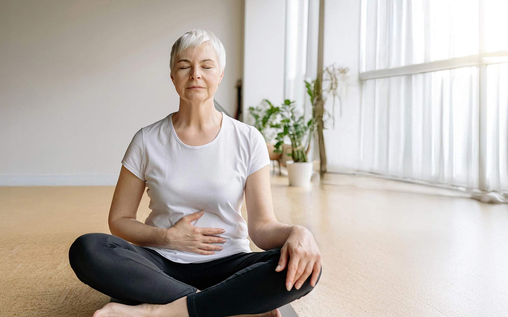 一位头发花白的老妇人盘腿坐在浅色地板上，练习正念或冥想以应对慢性疼痛。房间明亮，充满自然光，背景中有大窗户和室内植物。