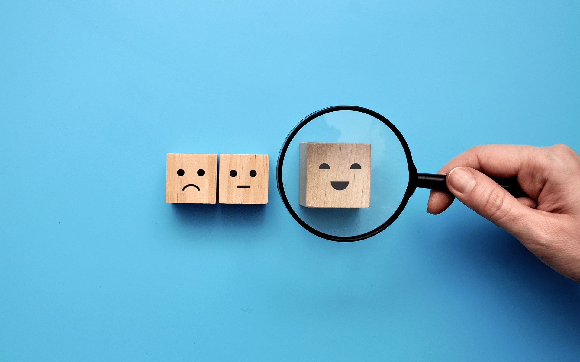 The Power of Leading with Appreciative Joy - Image of three wooden blocks on a blue background with facial expression drawn on them. A hand holds a magnifying glass over the one with a happy face.