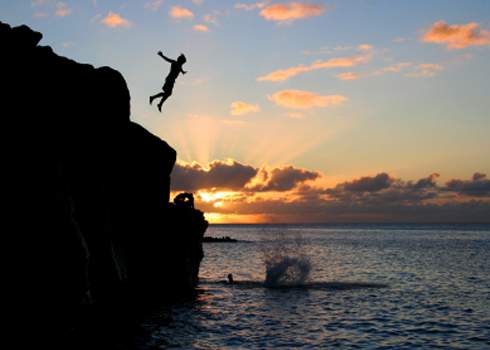一个人在夕阳下跳入海中。天空充满橙色和粉色的色彩，云层间露出夕阳。水下的溅起表明有人已经跳下去。场景在五彩斑斓的天空下显得非常美丽。