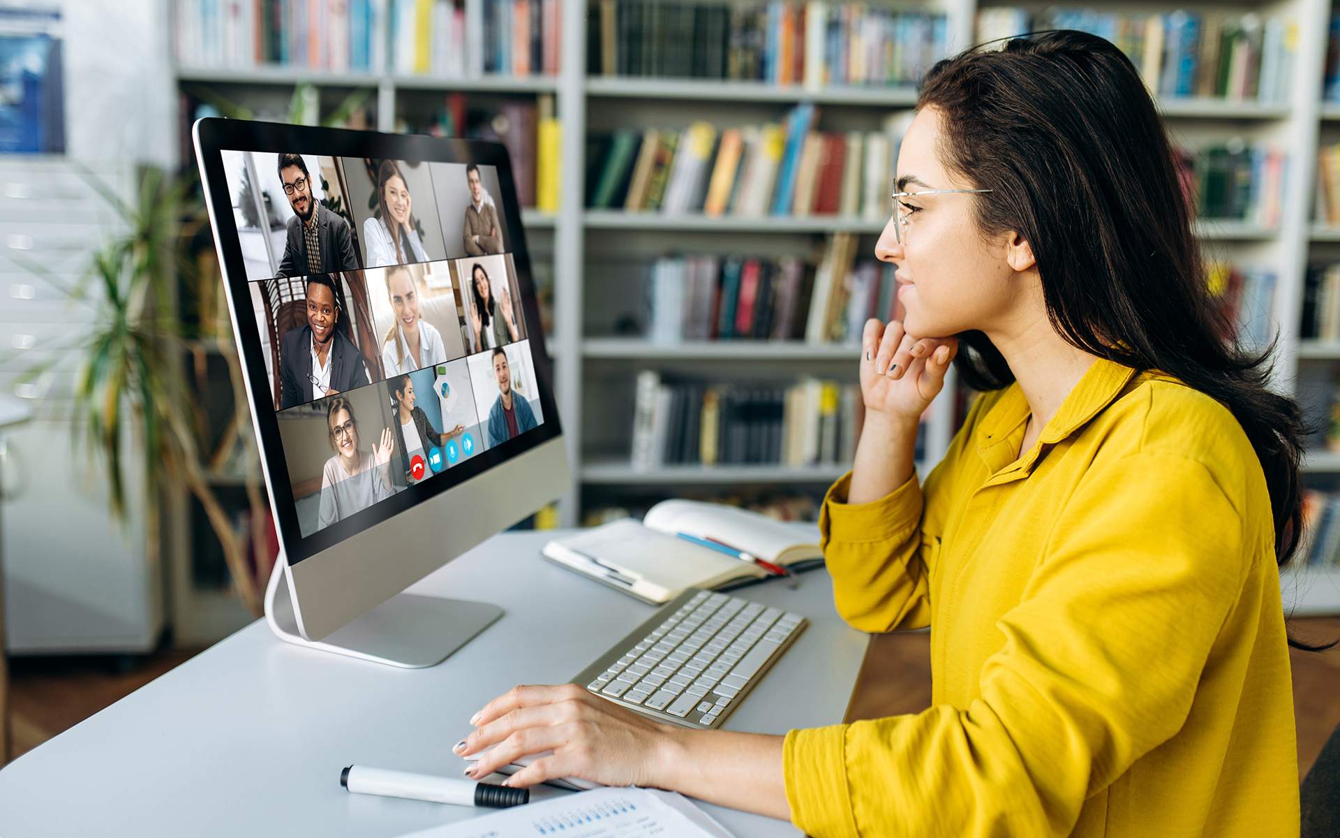 一位穿着黄色毛衣的女士坐在办公桌前与九个人进行视频通话。